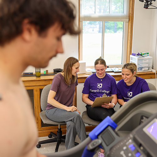 Student in the foreground on a treadmill, with students and faculty member discussing research data in the background.