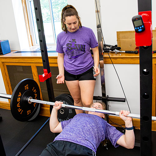 Student lifting barbells from a prone position with another student acting as spotter behind her.