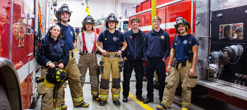 7 Cornell students in fire and EMT gear next to fire trucks