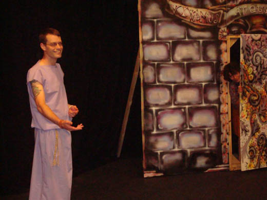 a smiling man gestures to something out of view, he wears a long purple dress. nearby someone is slipping out of the door while being careful to keep an eye on the man. 