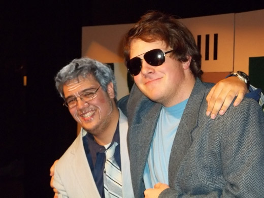 two men are shown from the chest up, they stand with their arms around each other, facing the camera. one wears sunglasses, a grey suit jacket, and a blue t-shirt, he smiles slightly. the other man, made to look older with makeup, wears a white suit, blue button-up shirt, and a striped tie. he smiles broadly. behind them you can see part of the set for the play. 