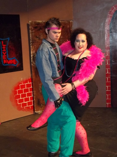 a woman in a low-cut black dress, pink feather boa, jewelry, and pink heart-printed hosier, has her arm around a man and has her leg up around his thigh. she's smiling. the man has his arm around her and his other hand holds her leg. he wears a pink workout headband, jean jacket, and turquoise pants. he looks confused. behind them you can see part of the set, parts are painted to look like brick and another part is painted to look like a neon sign saying "LIVE NUDE" 