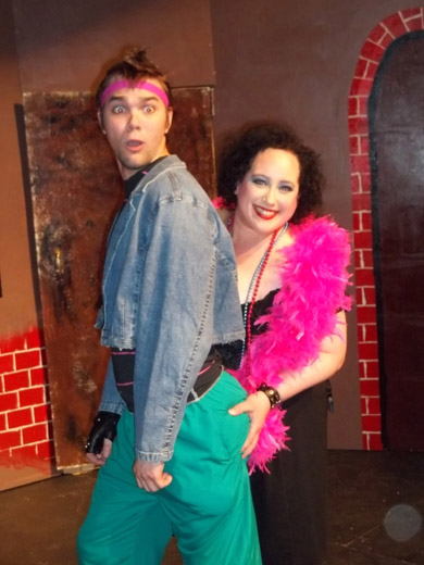 a smiling woman grabs a man's butt, he looks shocked. the woman wears a black dress and pink feather boa, the man is wearing a jean jacket, pink workout headband, and turquoise pants. 