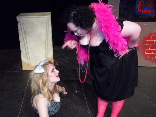 a woman with mussed hair and a large white bow sits on the florr. she wears a plaid, low-cut dress. she looks up indignantly at another woman scolding her. the other woman wears a black, low-cut dress, pink feather boa, jewelry, and pink hosiery. 