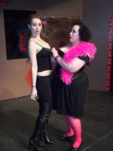 a woman in a tight tank top and black skinny jeans, exposing her midriff, as well as necklaces and thigh-high black boots, looks at the camera gobsmacked. another woman holds her by the front neckline of her tank top, and looks at her sternly. she is wearing a low-cut black dress and a pink feather boa