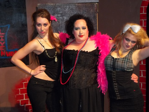 three women stand together, posing for the camera. they have their arms around one another. on the left is a woman with her hand on her hip. she wears a tight tank top and black skinny jeans as well as a couple necklaces. in the middle is a woman wearing a low-cut black dress with a black corset overtop and a feather boa, she smiles slightly. on the right is a woman with a large white bow in her hair, she has her arm up on the back of her head. she wears large hoop earrings, a plaid and black corset and a tight, black skirt 