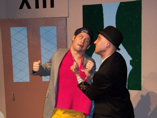 two men are in close quarters with each other about to fight. one wears a pink shit, yellow pants, a grey suit jacket, blue bandana, and a long gold chain. he has his fist raised and pulled back as if he's about to punch  the other man who is holding onto him and pulling him by the front of his shirt as well as his necklace. the other man wears a black suit and a bowler hat. 