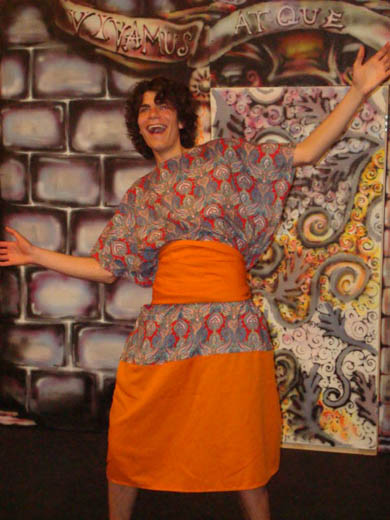 a man stands very excitedly, smiling largely, and with his arms outstretched. he wears a dress of orange as well as an intricate pattern in blue and red