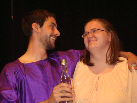 a man and a woman look fondly at each other. the man has his arm around her shoulders and wears purple, the woman wears white. the man is also holding a partially empty bottle.