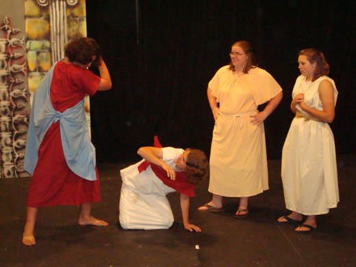 one man has clearly juist stricken anothe man who has fallen to his hands and knees on the floor. the man standing wears red and blue, the man stricken wears white and red and has one arm up to shield his face from further attacks. two women stand nearby watching, they wear white dresses