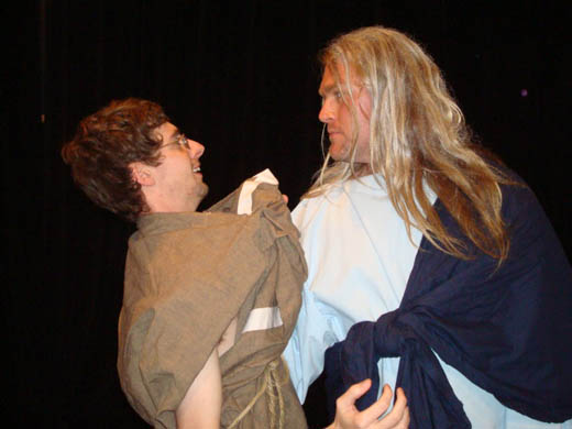 a man with long blond hair, wearing white and dark blue, threateningly holds onto the front of a brown tunic worn by another man who seems to be smiling in order to placate the other man. 