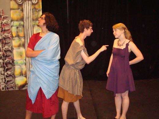 a man wearing  long, brown roman style dress, scolds a woman. the woman stands his her hand on her back looking unimpressed. she wears a statement necklace and a purple dress. next to them is another man, turning his back and his nose up at them. he wears a long, red tunic and a light blue sheet over his shoulder