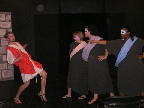 a man wearing sandals and a long white tunic with a bright red vest over it, leans back as if having been pushed. three people wearing long, dark grey dresses, look accusingly at him. the leftmost has a pink mask and a pink sash, the one in the middle has a purple mask and sash, the rightmost has a blue mask and sash and is pointing the the man. 