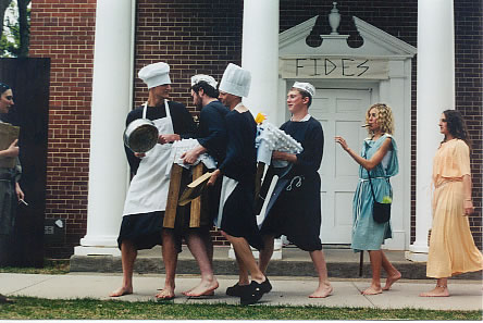 four men dressed as cooks are walking with pans and carrying chairs dressed up to look like a sheep. behind them are two women in long dresses. one has a bag slung over her shoulder and is playing a flute, the other holds her flute at her side.