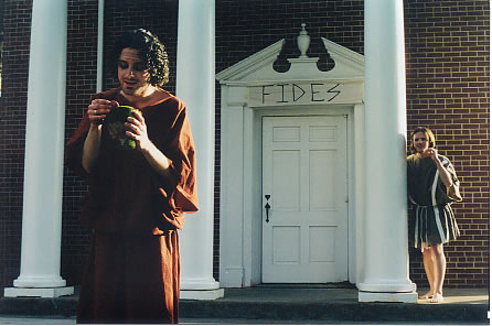 A man wearing a long red tunic tied in at the hips, faces away from a building and hold a large cup of gold coins, he admires them. behind him a man watches on, leaning against a column of the building, and wears a shorter grey tunic.