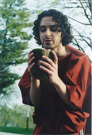 a man in a large, red tunic holds a large cup of gold coins close to his face and appears to be speaking to them