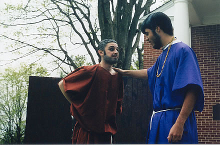 a man made to look old, wears a red tunic tied in at the hips, his arms are behind his back. another man wearing a blue tunic tied in at the waist, wears a gold chain and has his hand on the other man's shoulder