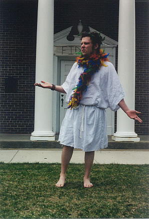 A barefoot man stands in front of a building with his arms out questioningly. he wears a knee-length tunic tied in at the waist, a colorful feather boa around his neck, and a wreath on his head.