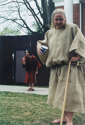 Outside, a woman stands barefoot in the foreground with a wooden cane. she wears a beige, ankle-length tunic, ties in at the hips. she holds a pepsi cup. behind her, a man stands pointing disapprovingly at something out of the frame. he wears a knee-length, red tunic, tied in at the hips