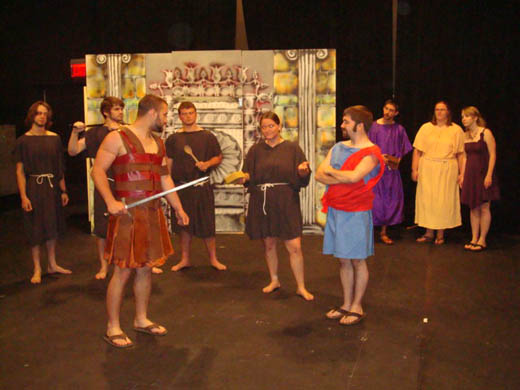 a man in "leather" armor holds a sword and faces a woman who has a sponge in one hand and is shrugging. three other people dressed as cooks stand in line with her and on her other side is a man with his arms crossed wearing red and blue. behind them a man wearing purple, a woman wearing white, and a woman wearing purple, look on