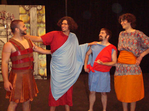 four men stand and smile at each other. from left to right is a man wearing "leather" armor and a dagger clapping the man next to him on the back, the next man has a long red tunic on under a wide blue sash, he has his hands on the shoulders of the man standing beside him. the next man wears a knee-length blue tunic with a wide red sash overtop and gestures to the first man. the last man has his hands on his hips and smiles knowingly, he wears a dress of orange and an intricate pattern in blue and red. 