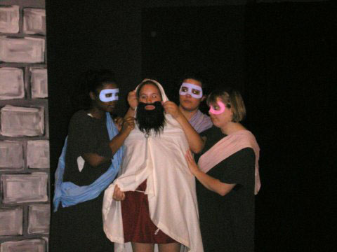 three people stand around another person wearing a beard. the three people have dark robes and colored masks and sashes. they are putting a white robe over the person in the middle of them who has a red tunic underneath