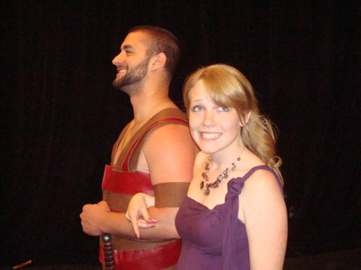 a woman wearing a purple dress smiles at something out of view, she holds the arm of a man wearing "leather" armor who is also smiling, but he looks directly ahead.