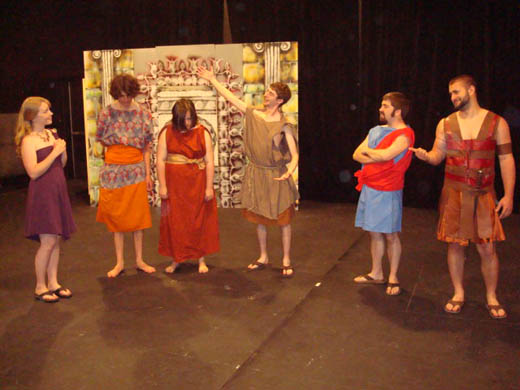 a man presents two people, who stand with their heads down, to a woman wearing a purple dress. the two people are barefoot and wear a blue patterned, orange and red dresses. the man presenting wears brown. two men look on, one wearing blue and red and the other wearing "leather" armor who points with his thumb at one of the others while appearing to say something funny to the man standing next to him.