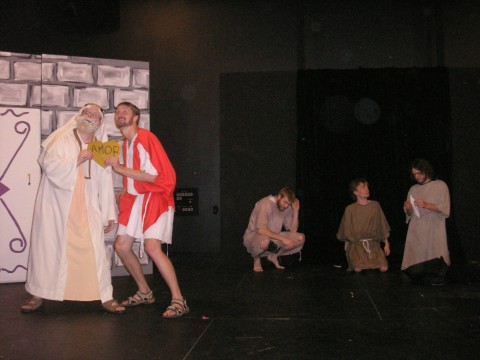 a man wearing a white beard and and white robes stands next to another man who appears much younger and who is wearing a long, white tunic with a red vest overtop. they hold a small hearty shaped sign that says "AMOR." Behind them three people in grey and brown robes crouch or kneel on the floor.