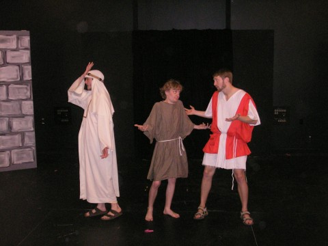 a man turned away from two others has his hand to his forehead in exasperation, he wears a long white dress and a head covering. next to him are two men, one wearing a brown tunic and the other wearing a white and red tunic. they look at each other shrugging 