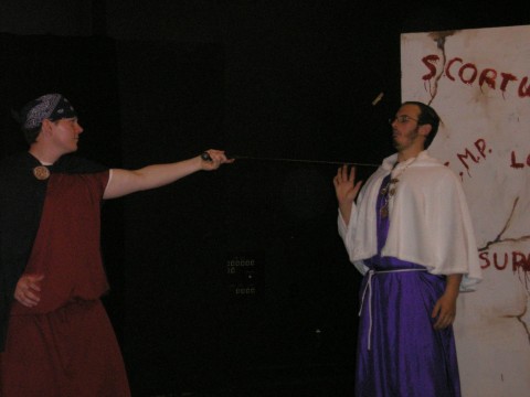 a man with a fighting stance has his arm outstretched pointing a sword at another man backed up against a wall. the first man has a bandana and a dark red tunic, the second man is wearing a purple tunic with a white elbow length cape overtop and a statement necklace. the wall behind him has large words that appear to have been written with blood