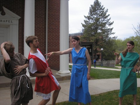 two men lean back as if having been pushed, one, standing directly behind the other, wears a grey tunic. the other wears a white tunic with a red vest overtop. opposite them are two women. one points at them and wears a long blue dress. the other, standing off a bit, strikes a sassy pose