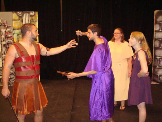 a man wearing "leather" armor, reaches out for a necklace held tightly by another man wearing purple. the second man seems to be pulling them back from the other guy. a woman in purple stands behind the second man smiling with her hands on her hips. further behind them is another woman wearing a long white dress.